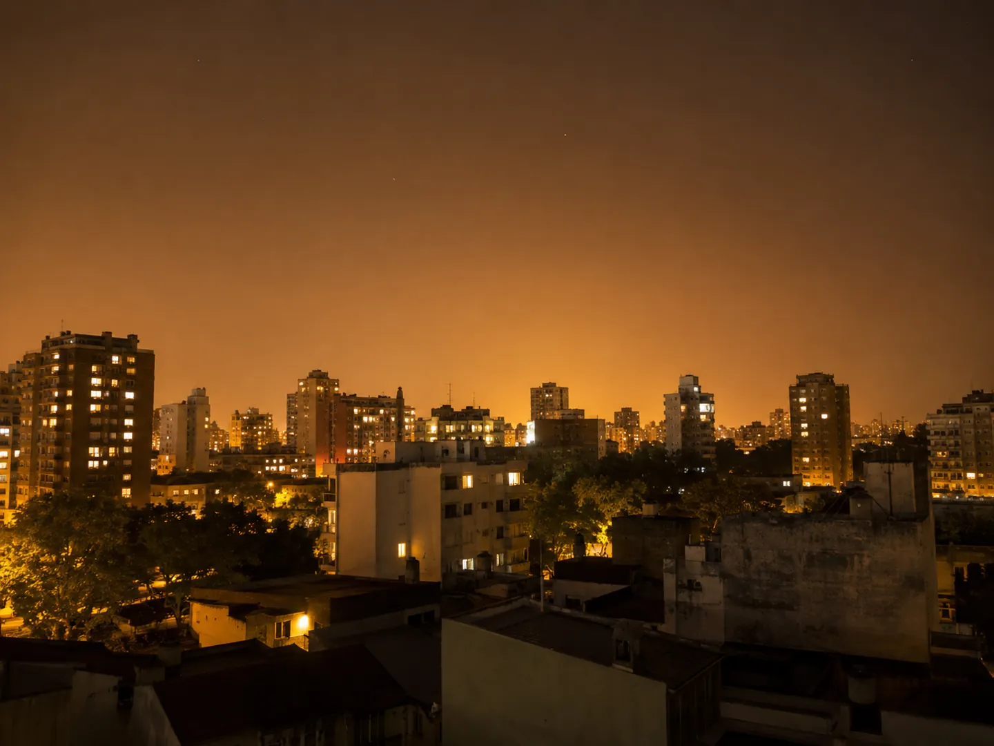 A city skyline of apartment blocks at night under a thick orange glow, with almost no stars visible above the rooftops