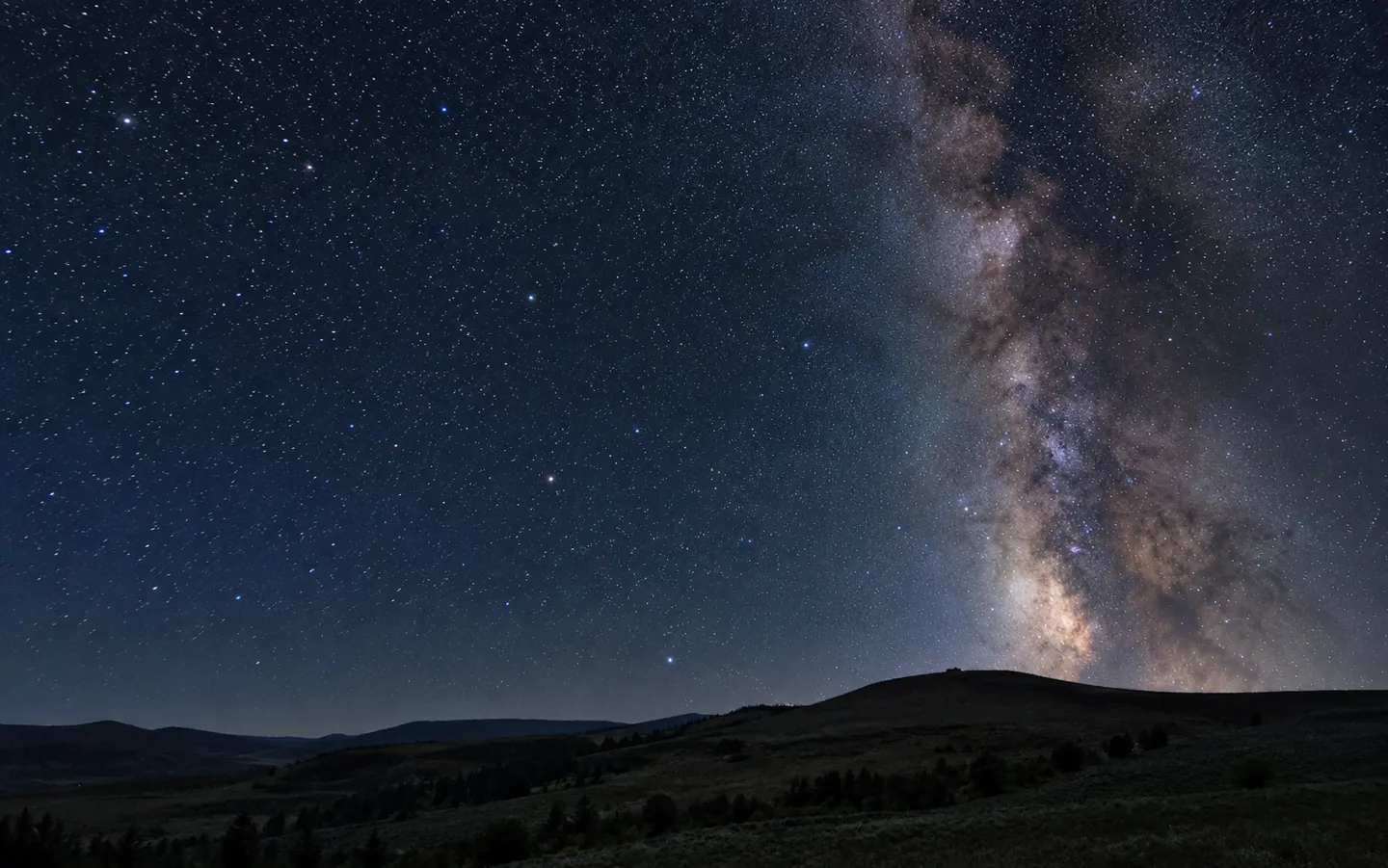 A clear night sky packed with stars above a dark rolling hillside, with the band of the Milky Way rising on the right